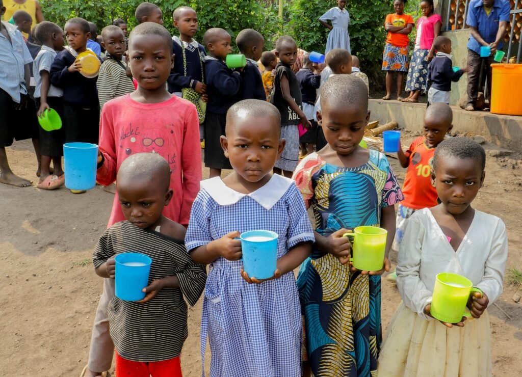 Group of children holding cups, standing in line outdoors, in a rural setting, waiting for food distribution.